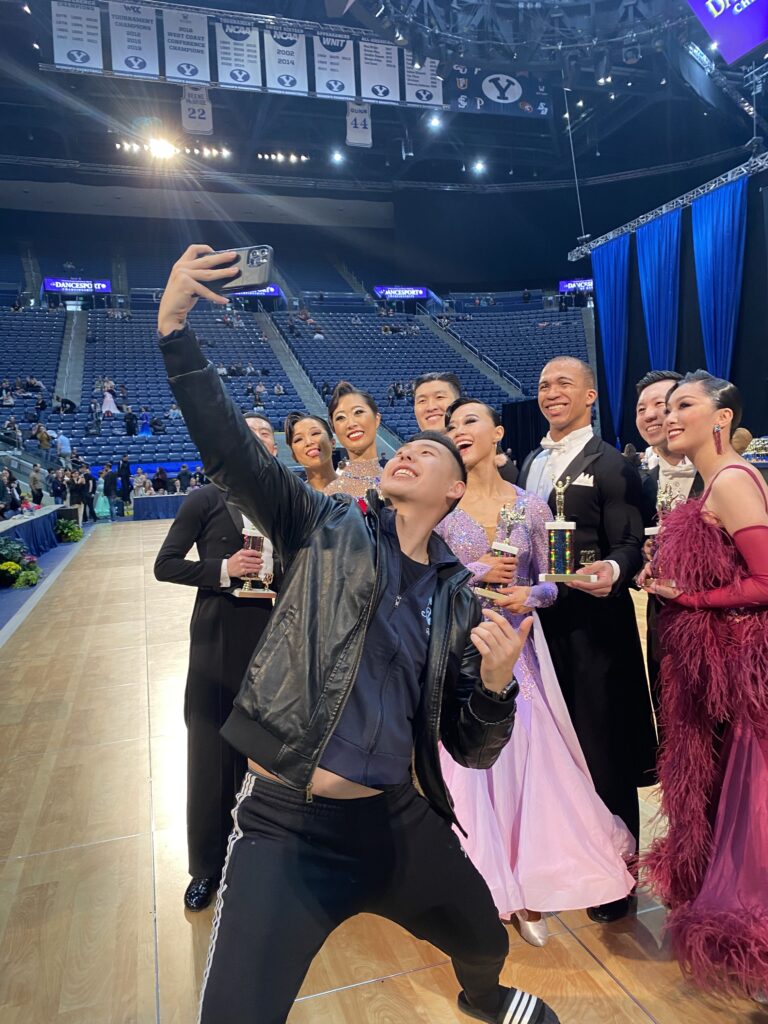 A group of nine collegiate ballroom dancers, eight of them Asian American and one of them Black, gathered for a group picture at a competition.