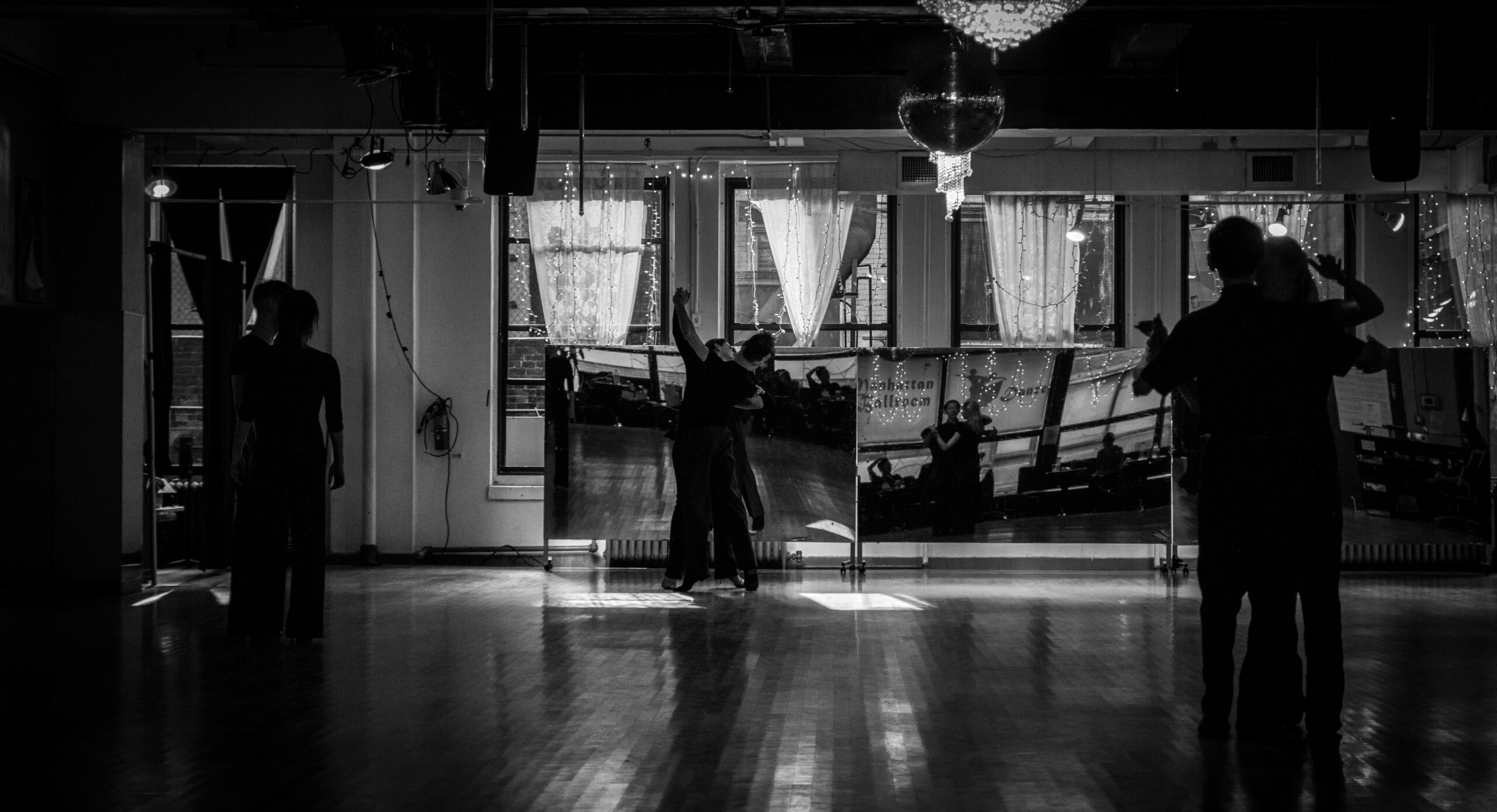 Several pairs of ballroom dancers practicing in a large mirrored studio, some moving in dance frame and some standing in place.